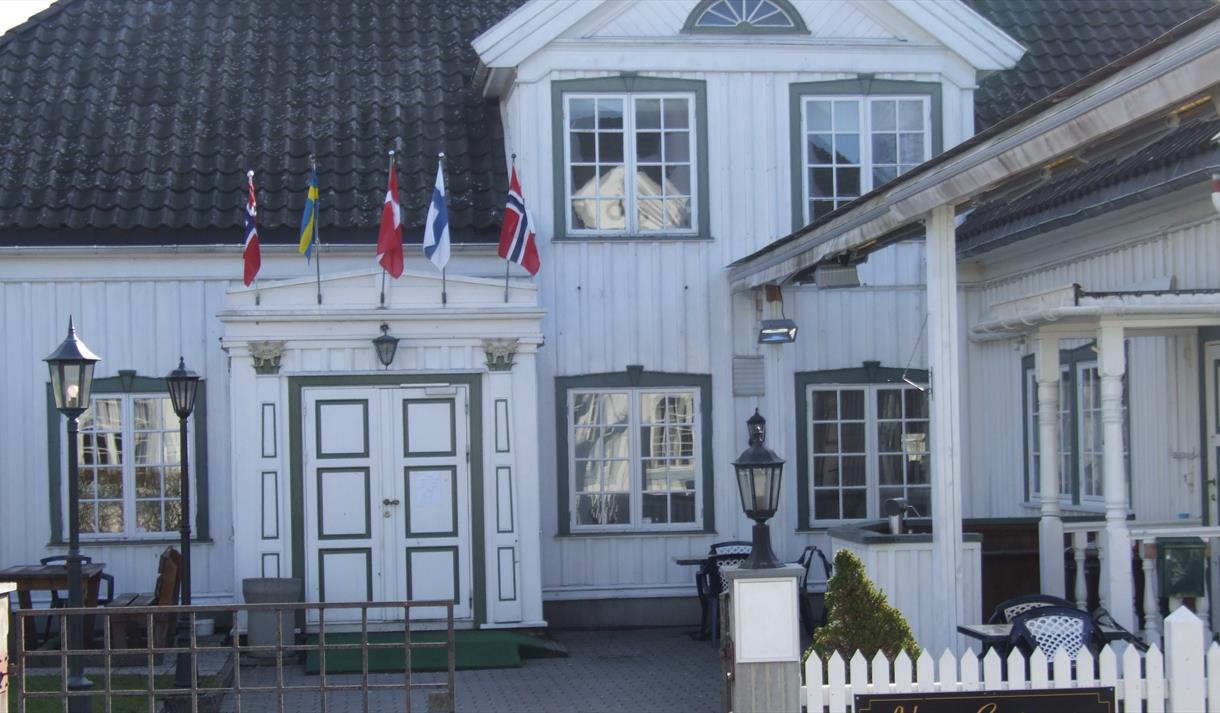 gammel hvit trebygning med blå vinduskarmer, smijernsport og flagg