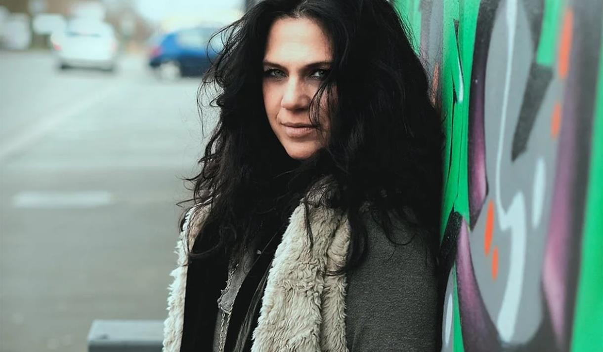 dame med sort langt hår stående inntil en vegg med grafitti