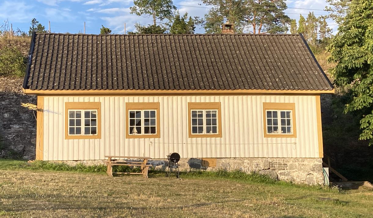gammelt beige trehus med oransje karmer rundt smårutete vinduer. Gressplen med trær i bakgrunnen og sittebenk