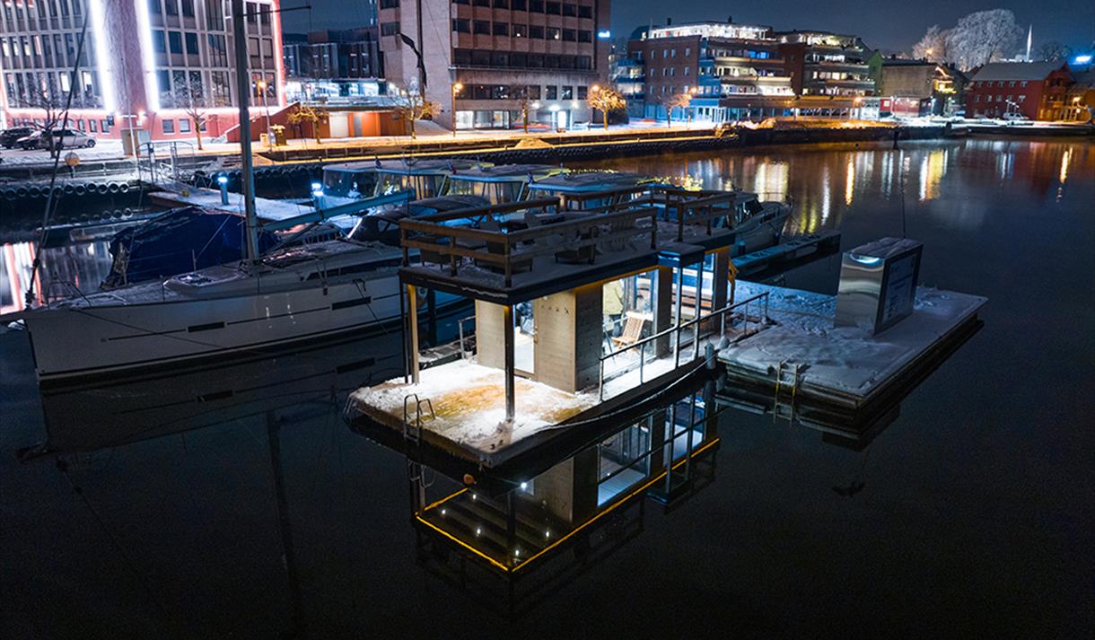 sauna liggene ved elva, med båter på brygge. blokker med lys i bakgrunnen